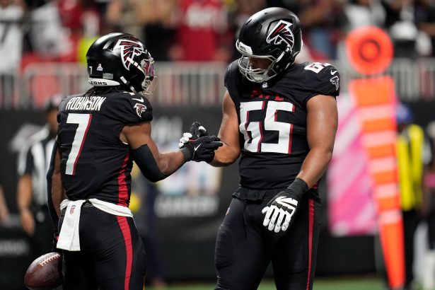 Zwei Footballspieler der Atlanta Falcons in schwarzen Trikots feiern auf dem Spielfeld in Berlin. Einer von ihnen hält einen Football in der Hand und reicht seinem Teamkollegen von der Offensive Line die Hand zum Handschlag. Im Hintergrund sind unscharfe Fans, Seitenlinienmarkierungen und Stadionlichter zu sehen. Diese Beschreibung wurde mit der FootballR KI automatisch generiert.