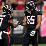 Zwei Footballspieler der Atlanta Falcons in schwarzen Trikots feiern auf dem Spielfeld in Berlin. Einer von ihnen hält einen Football in der Hand und reicht seinem Teamkollegen von der Offensive Line die Hand zum Handschlag. Im Hintergrund sind unscharfe Fans, Seitenlinienmarkierungen und Stadionlichter zu sehen. Diese Beschreibung wurde mit der FootballR KI automatisch generiert.