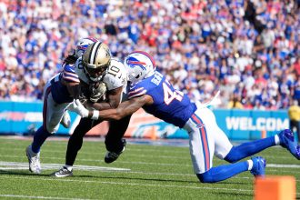 Zwei Verteidiger der Buffalo Bills in blauen Uniformen greifen den Saints-Receiver Brandin Cooks an, der den Football während eines NFL-Spiels in der Hand hält. Die Szene spielt auf einem sonnenbeschienenen Feld mit einer unscharfen Menschenmenge im Stadionhintergrund. Diese Beschreibung wurde mit der FootballR KI automatisch generiert.