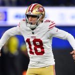 Eddy Piñeiro - Ein Football-Spieler der San Francisco 49ers in einem weißen Trikot (Nummer 18), einer goldenen Hose und einem goldenen Helm mit rotem Streifen jubelt auf dem Spielfeld mit angewinkelten Armen und offenem Mund, während im Hintergrund unscharfe Zuschauer jubeln. Diese Beschreibung wurde mit der FootballR KI automatisch generiert.