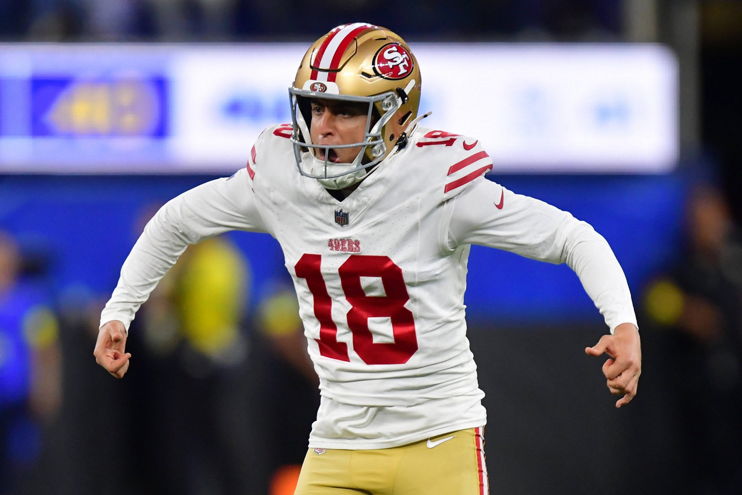 Eddy Piñeiro - Ein Football-Spieler der San Francisco 49ers in einem weißen Trikot (Nummer 18), einer goldenen Hose und einem goldenen Helm mit rotem Streifen jubelt auf dem Spielfeld mit angewinkelten Armen und offenem Mund, während im Hintergrund unscharfe Zuschauer jubeln. Diese Beschreibung wurde mit der FootballR KI automatisch generiert.