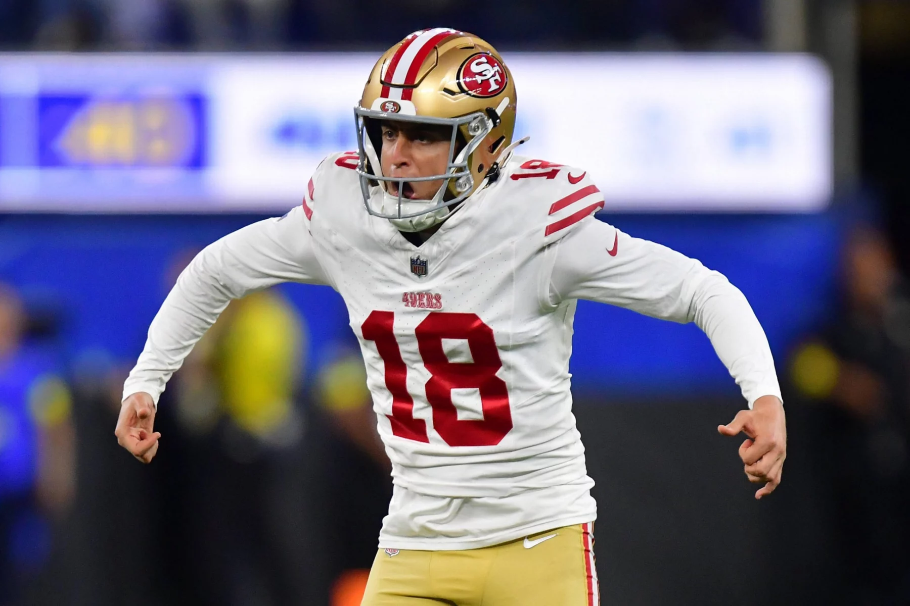 Eddy Piñeiro - Ein Football-Spieler der San Francisco 49ers in einem weißen Trikot (Nummer 18), einer goldenen Hose und einem goldenen Helm mit rotem Streifen jubelt auf dem Spielfeld mit angewinkelten Armen und offenem Mund, während im Hintergrund unscharfe Zuschauer jubeln. Diese Beschreibung wurde mit der FootballR KI automatisch generiert.