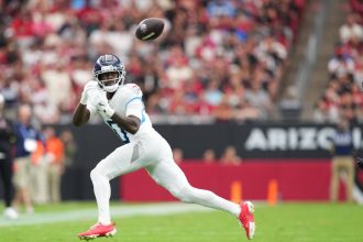 Calvin Ridley - Ein Top-Receiver in einer weiß-blauen Titans-Uniform, mit roten Stollenschuhen und Handschuhen, bereitet sich darauf vor, einen Football in der Luft auf einem grünen Feld zu fangen. Das Stadion ist gefüllt mit verschwommenen Zuschauern und einem schwarzen Banner mit "ARIZO", das teilweise hinter ihm zu sehen ist. Diese Beschreibung wurde mit der FootballR KI automatisch generiert.