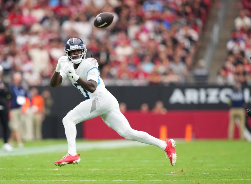Calvin Ridley - Ein Top-Receiver in einer weiß-blauen Titans-Uniform, mit roten Stollenschuhen und Handschuhen, bereitet sich darauf vor, einen Football in der Luft auf einem grünen Feld zu fangen. Das Stadion ist gefüllt mit verschwommenen Zuschauern und einem schwarzen Banner mit "ARIZO", das teilweise hinter ihm zu sehen ist. Diese Beschreibung wurde mit der FootballR KI automatisch generiert.