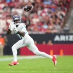 Calvin Ridley - Ein Top-Receiver in einer weiß-blauen Titans-Uniform, mit roten Stollenschuhen und Handschuhen, bereitet sich darauf vor, einen Football in der Luft auf einem grünen Feld zu fangen. Das Stadion ist gefüllt mit verschwommenen Zuschauern und einem schwarzen Banner mit "ARIZO", das teilweise hinter ihm zu sehen ist. Diese Beschreibung wurde mit der FootballR KI automatisch generiert.