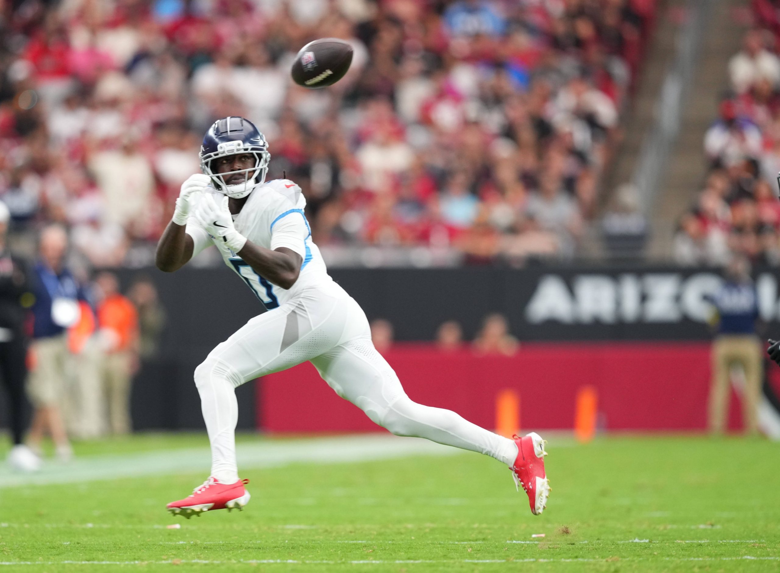 Calvin Ridley - Ein Top-Receiver in einer weiß-blauen Titans-Uniform, mit roten Stollenschuhen und Handschuhen, bereitet sich darauf vor, einen Football in der Luft auf einem grünen Feld zu fangen. Das Stadion ist gefüllt mit verschwommenen Zuschauern und einem schwarzen Banner mit "ARIZO", das teilweise hinter ihm zu sehen ist. Diese Beschreibung wurde mit der FootballR KI automatisch generiert.