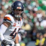 Alex Singleton - Ein Football-Spieler, der eine Denver Broncos-Uniform, einen Helm und ein Trikot mit der Nummer 49 trägt, steht während eines Spiels auf dem Spielfeld. Er hat lange Haare, einen entschlossenen Gesichtsausdruck und die OP-Initialen, die zur Unterstützung der Krebsbekämpfung angebracht sind. Diese Beschreibung wurde mit der FootballR KI automatisch generiert.