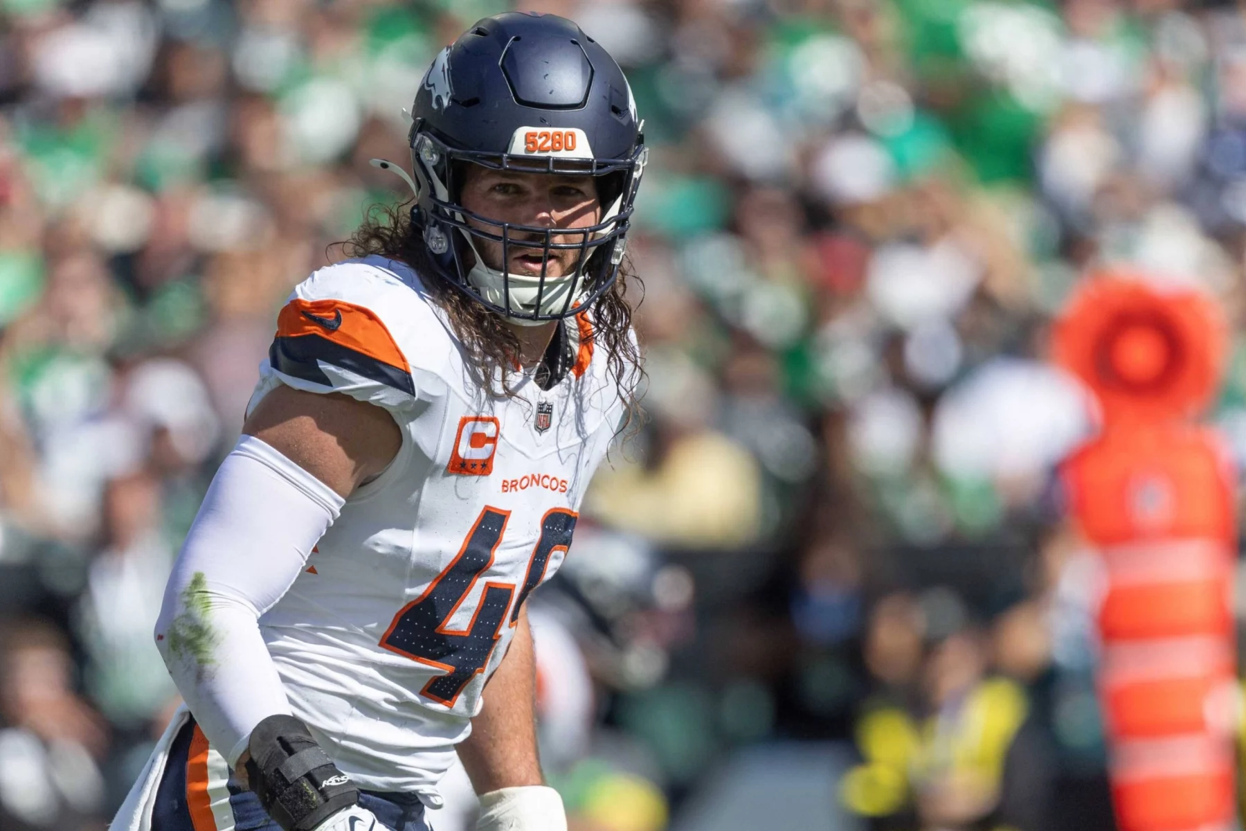 Alex Singleton - Ein Football-Spieler, der eine Denver Broncos-Uniform, einen Helm und ein Trikot mit der Nummer 49 trägt, steht während eines Spiels auf dem Spielfeld. Er hat lange Haare, einen entschlossenen Gesichtsausdruck und die OP-Initialen, die zur Unterstützung der Krebsbekämpfung angebracht sind. Diese Beschreibung wurde mit der FootballR KI automatisch generiert.