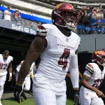 Frankie Luvu - Die Footballspieler der Washington Commanders in weißen Trikots, angeführt von Commanders-Linebacker Nummer 4 mit getöntem Visier, kommen aus einem Tunnel auf das Spielfeld des SoFi-Stadions. Im Hintergrund sind bei diesem NFL-Spiel Fans und Stadionplätze zu sehen. Diese Beschreibung wurde mit der FootballR KI automatisch generiert.