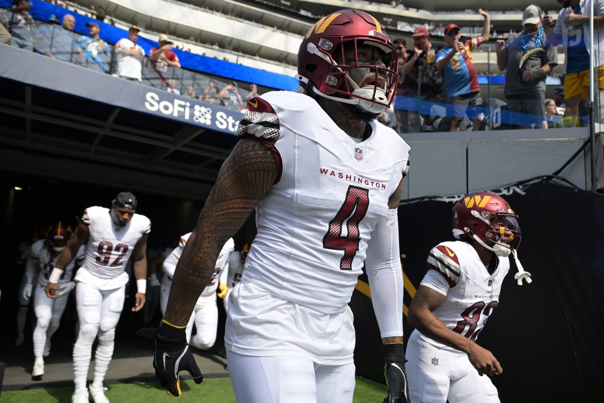 Frankie Luvu - Die Footballspieler der Washington Commanders in weißen Trikots, angeführt von Commanders-Linebacker Nummer 4 mit getöntem Visier, kommen aus einem Tunnel auf das Spielfeld des SoFi-Stadions. Im Hintergrund sind bei diesem NFL-Spiel Fans und Stadionplätze zu sehen. Diese Beschreibung wurde mit der FootballR KI automatisch generiert.