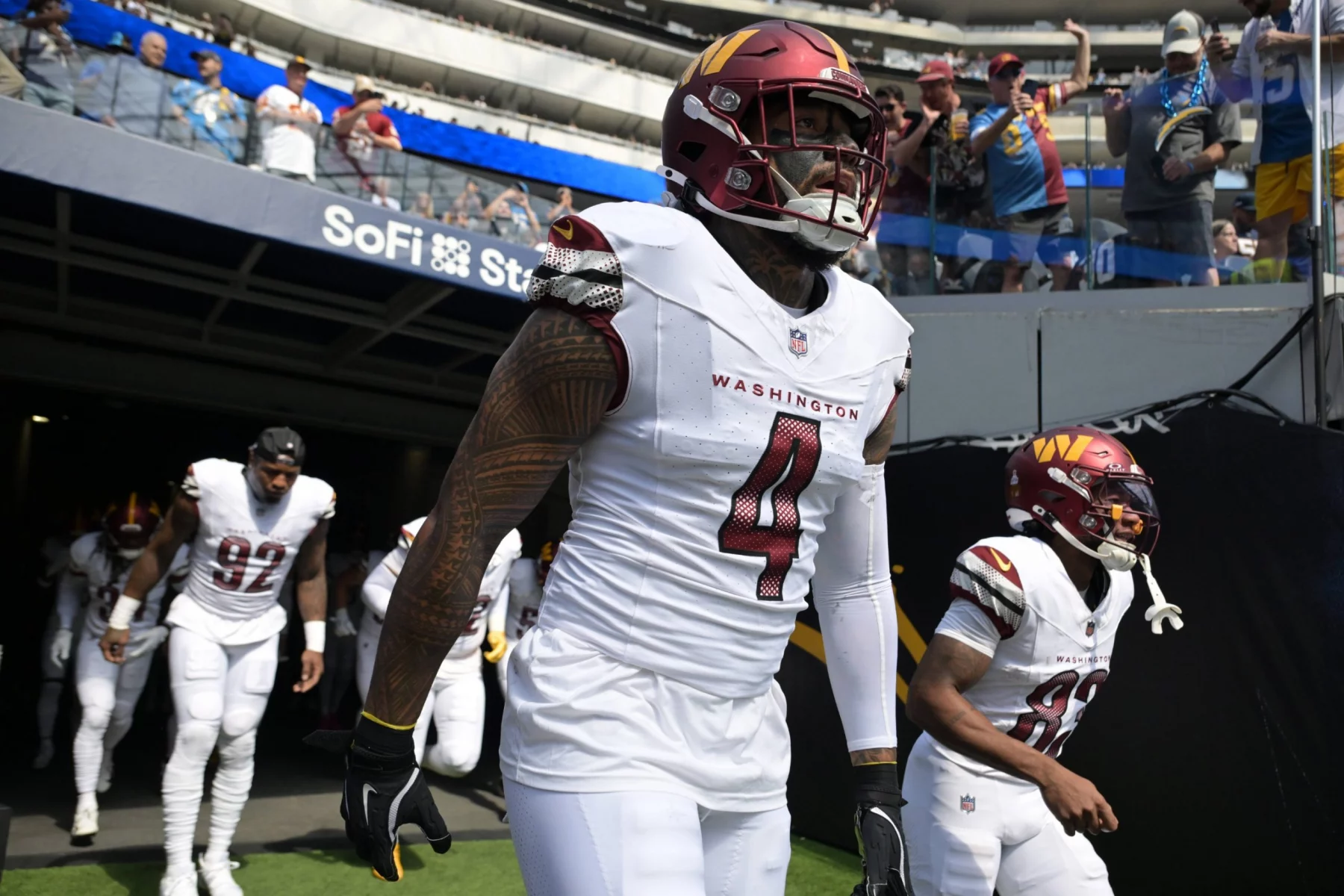 Frankie Luvu - Die Footballspieler der Washington Commanders in weißen Trikots, angeführt von Commanders-Linebacker Nummer 4 mit getöntem Visier, kommen aus einem Tunnel auf das Spielfeld des SoFi-Stadions. Im Hintergrund sind bei diesem NFL-Spiel Fans und Stadionplätze zu sehen. Diese Beschreibung wurde mit der FootballR KI automatisch generiert.