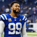 Ein Football-Spieler, DeForest Buckner, trägt ein blaues Indianapolis Colts-Trikot mit der Nummer 99 und steht auf dem Spielfeld. Er hat einen Bart und lächelt, der Stadionhintergrund ist unscharf, Menschen und Sitze sind zu sehen. Diese Beschreibung wurde mit der FootballR KI automatisch generiert.