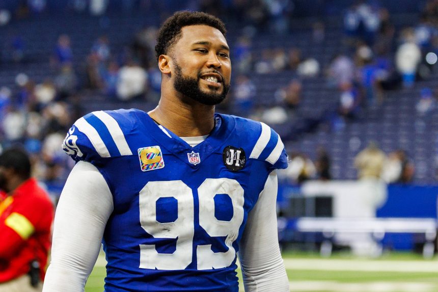Ein Football-Spieler, DeForest Buckner, trägt ein blaues Indianapolis Colts-Trikot mit der Nummer 99 und steht auf dem Spielfeld. Er hat einen Bart und lächelt, der Stadionhintergrund ist unscharf, Menschen und Sitze sind zu sehen. Diese Beschreibung wurde mit der FootballR KI automatisch generiert.