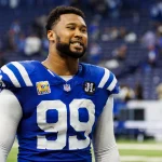 Ein Football-Spieler, DeForest Buckner, trägt ein blaues Indianapolis Colts-Trikot mit der Nummer 99 und steht auf dem Spielfeld. Er hat einen Bart und lächelt, der Stadionhintergrund ist unscharf, Menschen und Sitze sind zu sehen. Diese Beschreibung wurde mit der FootballR KI automatisch generiert.