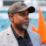 Ein Mann mittleren Alters mit grauem Bart, der dem Geschäftsführer der Dolphins, Chris Grier, ähnelt, trägt eine schwarze Mütze, einen grau karierten Blazer und ein schwarzes Hemd. Er blickt mit ernster Miene nach rechts. Hinter ihm befinden sich türkisfarbene Stadionsitze und ein orangefarbenes kronenähnliches Element. Diese Beschreibung wurde mit der FootballR KI automatisch generiert.