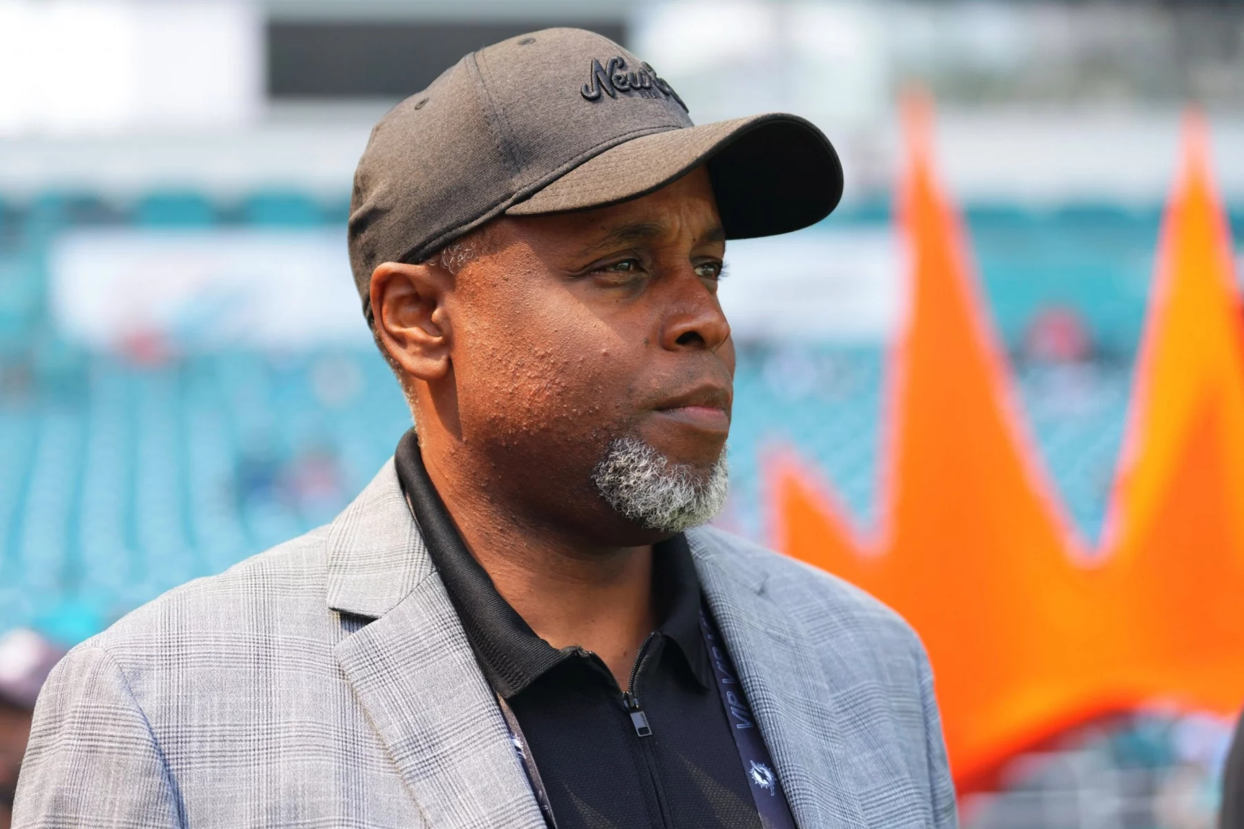 Ein Mann mittleren Alters mit grauem Bart, der dem Geschäftsführer der Dolphins, Chris Grier, ähnelt, trägt eine schwarze Mütze, einen grau karierten Blazer und ein schwarzes Hemd. Er blickt mit ernster Miene nach rechts. Hinter ihm befinden sich türkisfarbene Stadionsitze und ein orangefarbenes kronenähnliches Element. Diese Beschreibung wurde mit der FootballR KI automatisch generiert.