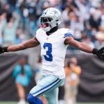 George Pickens der Dallas Cowboys mit der Trikotnummer 3 breitet seine Arme aus, während er einen Football hält und auf dem Spielfeld feiert. Er trägt einen silbernen Helm und eine weiße Uniform, während Cheerleader und Fans im Hintergrund des Cowboys-Stadions verschwimmen. Diese Beschreibung wurde mit der FootballR KI automatisch generiert.