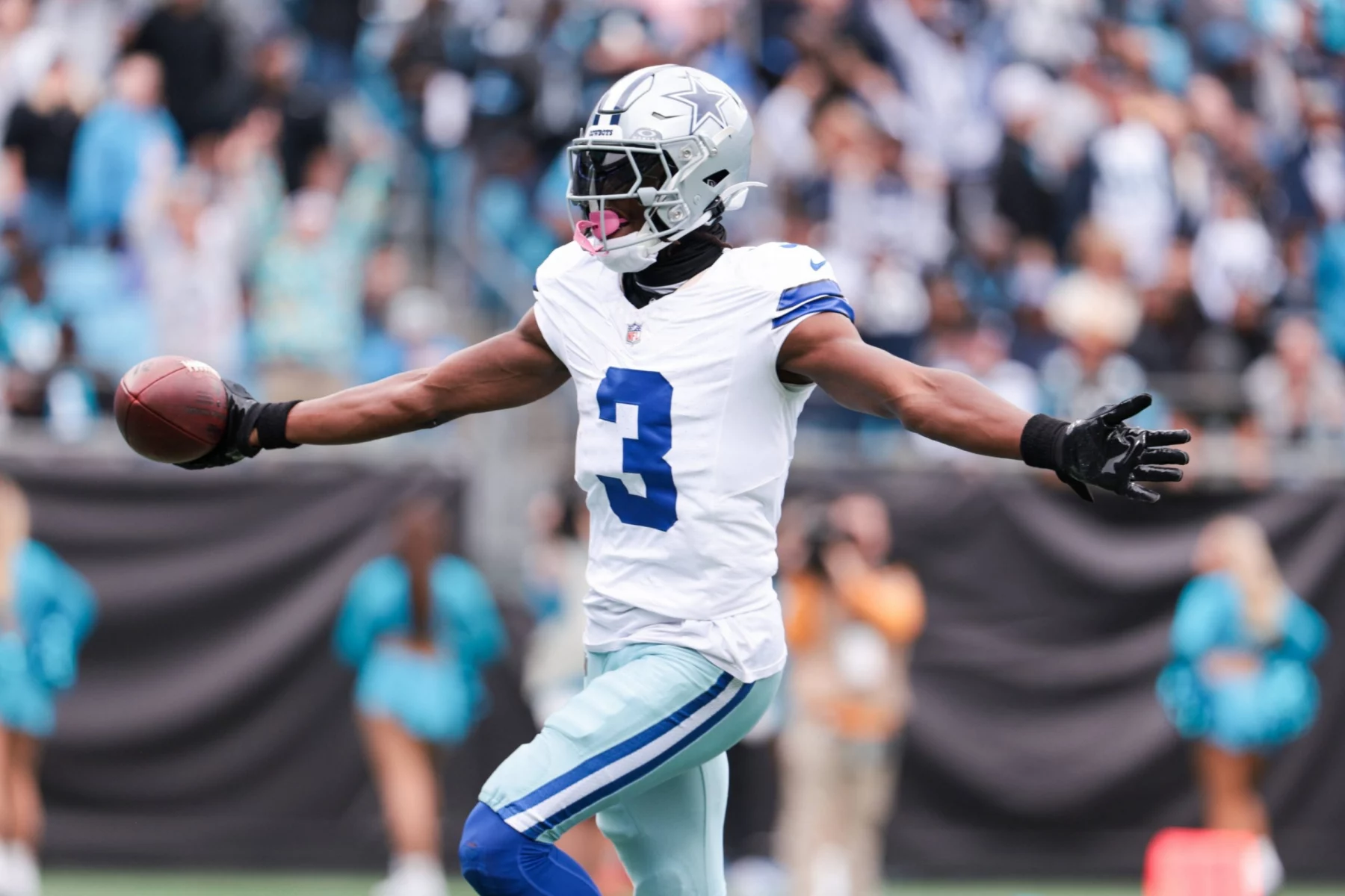 George Pickens der Dallas Cowboys mit der Trikotnummer 3 breitet seine Arme aus, während er einen Football hält und auf dem Spielfeld feiert. Er trägt einen silbernen Helm und eine weiße Uniform, während Cheerleader und Fans im Hintergrund des Cowboys-Stadions verschwimmen. Diese Beschreibung wurde mit der FootballR KI automatisch generiert.