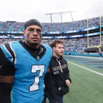 Tre'von Moehrig - Ein Footballspieler in einem blauen Panthers-Trikot mit der Nummer 7 geht mit seinem Helm in der Hand vom Spielfeld, begleitet von einem Mann in einem schwarzen Kapuzenpullover. Das Stadion ist mit Fans gefüllt und im Hintergrund sind Torpfosten zu sehen. Diese Beschreibung wurde mit der FootballR KI automatisch generiert.