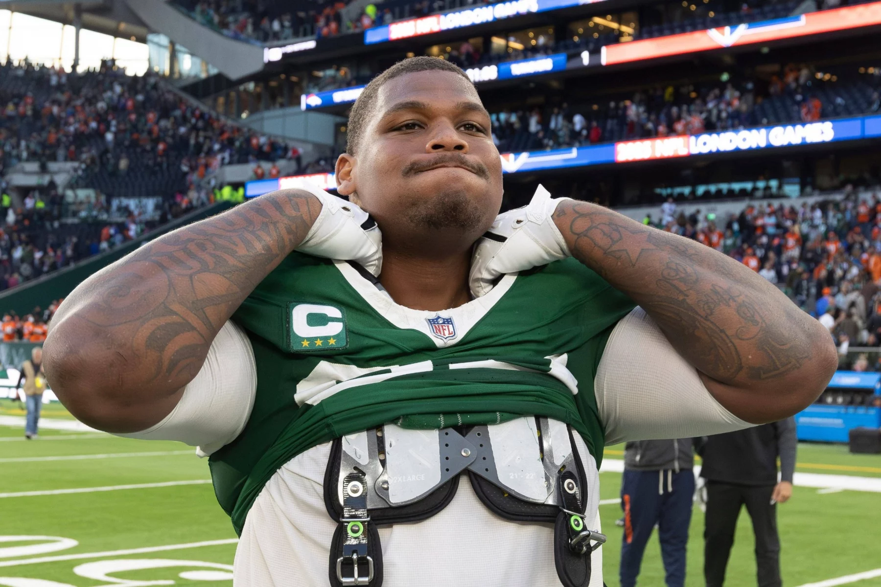 Quinnen Williams - Ein Footballspieler in einem grün-weißen Trikot passt seine Polster auf dem Spielfeld während der Handelsschlusswoche an. Er hat sichtbare Armtattoos, trägt ein Kapitänsabzeichen und steht vor einem vollen Stadion mit der Anzeigetafel und der Menge im Hintergrund. Diese Beschreibung wurde mit der FootballR KI automatisch generiert.