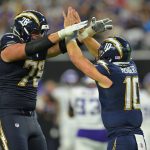 Zwei American-Football-Spieler in dunkelblauen Chargers-Trikots geben sich auf dem Spielfeld ein High-Five. Einer, der viel größer ist und die Nummer 76 trägt - Joe Alt - jubelt mit der Nummer 10, während im Hintergrund unscharfe Gegner in Weiß erscheinen. Diese Beschreibung wurde mit der FootballR KI automatisch generiert.