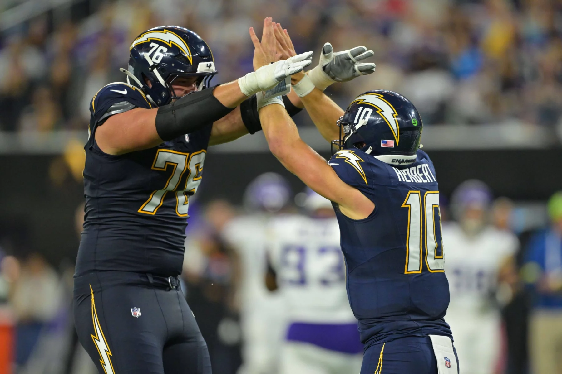 Zwei American-Football-Spieler in dunkelblauen Chargers-Trikots geben sich auf dem Spielfeld ein High-Five. Einer, der viel größer ist und die Nummer 76 trägt - Joe Alt - jubelt mit der Nummer 10, während im Hintergrund unscharfe Gegner in Weiß erscheinen. Diese Beschreibung wurde mit der FootballR KI automatisch generiert.