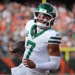 Justin Fields - Ein Football-Spieler in einem weiß-grünen Trikot der New York Jets, mit Helm und der Nummer 7, läuft mit dem Ball auf dem Spielfeld. Die unscharfe Menge und der Torpfosten erinnern an eine schnelle Bewegung während eines Spiels gegen die Cleveland Browns. Diese Beschreibung wurde mit der FootballR KI automatisch generiert.
