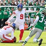 Der Kicker der Giants, Graham Gano, in weißer Uniform und roten Socken, kickt einen Football, der von einem Mannschaftskameraden gehalten wird, während ein Spieler der New York Jets (#34) auf sie zustürmt. Im Hintergrund sind Fans und Spieler zu sehen. Diese Beschreibung wurde mit der FootballR KI automatisch generiert.