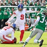 Der Kicker der Giants, Graham Gano, in weißer Uniform und roten Socken, kickt einen Football, der von einem Mannschaftskameraden gehalten wird, während ein Spieler der New York Jets (#34) auf sie zustürmt. Im Hintergrund sind Fans und Spieler zu sehen. Diese Beschreibung wurde mit der FootballR KI automatisch generiert.