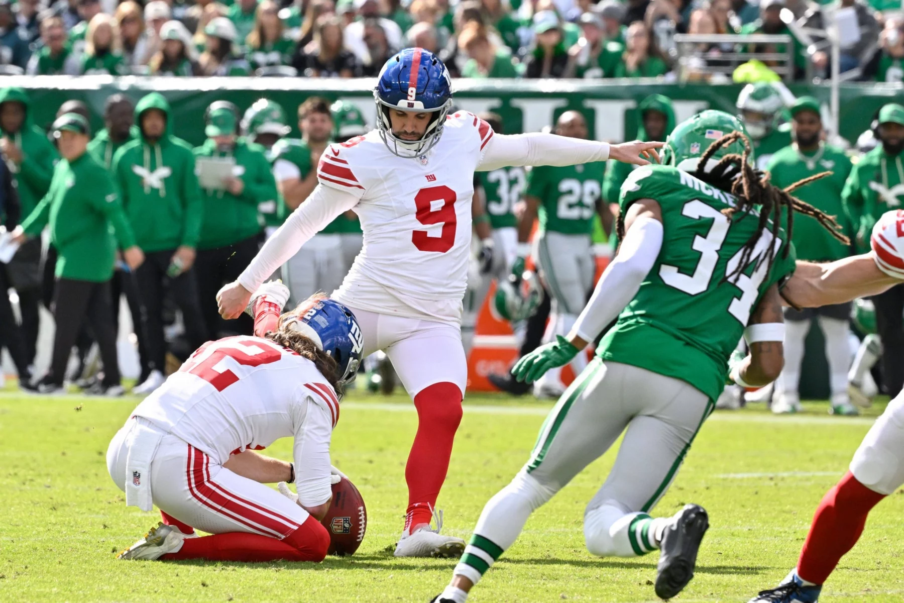 Der Kicker der Giants, Graham Gano, in weißer Uniform und roten Socken, kickt einen Football, der von einem Mannschaftskameraden gehalten wird, während ein Spieler der New York Jets (#34) auf sie zustürmt. Im Hintergrund sind Fans und Spieler zu sehen. Diese Beschreibung wurde mit der FootballR KI automatisch generiert.