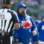 Der Trainer der New York Giants, Brian Daboll, trägt einen blauen Kapuzenpulli und eine Mütze, trägt ein Headset und schreit, während er eine rote Fahne mit der Aufschrift "DJ 13" in Richtung eines Schiedsrichters hält. Im unscharfen Hintergrund sind andere Teammitglieder und die Zuschauer zu sehen. Diese Beschreibung wurde mit der FootballR KI automatisch generiert.