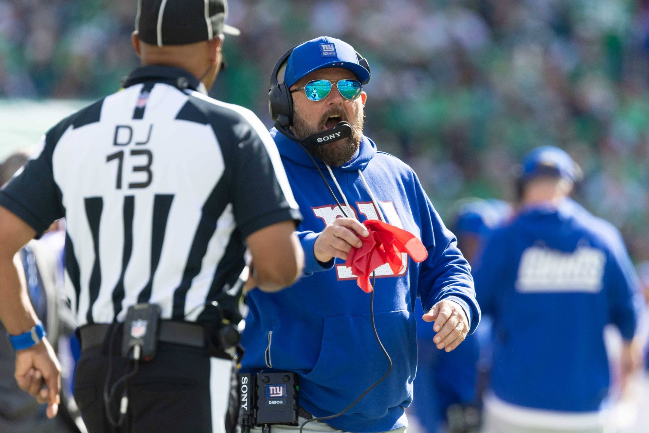 Der Trainer der New York Giants, Brian Daboll, trägt einen blauen Kapuzenpulli und eine Mütze, trägt ein Headset und schreit, während er eine rote Fahne mit der Aufschrift "DJ 13" in Richtung eines Schiedsrichters hält. Im unscharfen Hintergrund sind andere Teammitglieder und die Zuschauer zu sehen. Diese Beschreibung wurde mit der FootballR KI automatisch generiert.