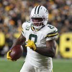 Ein Packers-Footballspieler, Nummer 9, sprintet während eines Thanksgiving-Spiels mit dem Ball. Er trägt gelbe Handschuhe, einen Helm mit dunklem Visier und wird von einer verschwommenen Menge angefeuert, während ein Schiedsrichter in der Nähe zusieht. Josh Jacobs sticht in den Farben von Green Bay hervor. Diese Beschreibung wurde mit der FootballR KI automatisch generiert.