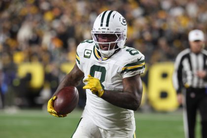 Ein Packers-Footballspieler, Nummer 9, sprintet während eines Thanksgiving-Spiels mit dem Ball. Er trägt gelbe Handschuhe, einen Helm mit dunklem Visier und wird von einer verschwommenen Menge angefeuert, während ein Schiedsrichter in der Nähe zusieht. Josh Jacobs sticht in den Farben von Green Bay hervor. Diese Beschreibung wurde mit der FootballR KI automatisch generiert.