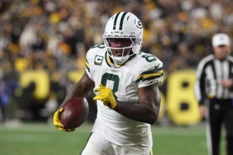 Ein Packers-Footballspieler, Nummer 9, sprintet während eines Thanksgiving-Spiels mit dem Ball. Er trägt gelbe Handschuhe, einen Helm mit dunklem Visier und wird von einer verschwommenen Menge angefeuert, während ein Schiedsrichter in der Nähe zusieht. Josh Jacobs sticht in den Farben von Green Bay hervor. Diese Beschreibung wurde mit der FootballR KI automatisch generiert.