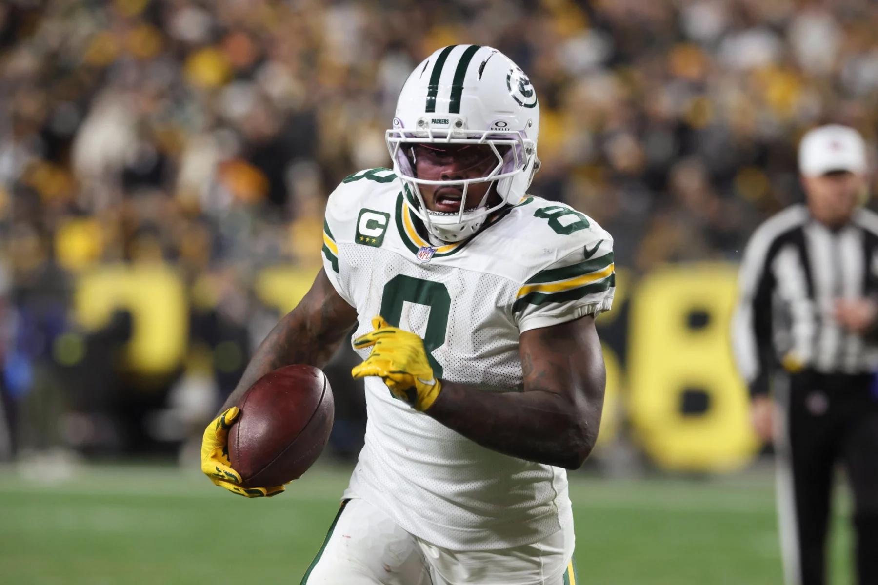 Ein Packers-Footballspieler, Nummer 9, sprintet während eines Thanksgiving-Spiels mit dem Ball. Er trägt gelbe Handschuhe, einen Helm mit dunklem Visier und wird von einer verschwommenen Menge angefeuert, während ein Schiedsrichter in der Nähe zusieht. Josh Jacobs sticht in den Farben von Green Bay hervor. Diese Beschreibung wurde mit der FootballR KI automatisch generiert.