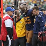 Tucker Kraft - Ein Footballspieler mit braunem Helm und marineblauem Trikot, bei dem der Verdacht auf einen Kreuzbandriss besteht, wird von zwei Mitarbeitern vom Spielfeld getragen. Andere Mitarbeiter, die Jacken und Mützen tragen, stehen in der Nähe. Im Hintergrund sind die Zuschauer und die Seitenlinie zu sehen. Diese Beschreibung wurde mit der FootballR KI automatisch generiert.