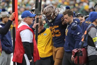 Tucker Kraft - Ein Footballspieler mit braunem Helm und marineblauem Trikot, bei dem der Verdacht auf einen Kreuzbandriss besteht, wird von zwei Mitarbeitern vom Spielfeld getragen. Andere Mitarbeiter, die Jacken und Mützen tragen, stehen in der Nähe. Im Hintergrund sind die Zuschauer und die Seitenlinie zu sehen. Diese Beschreibung wurde mit der FootballR KI automatisch generiert.