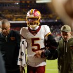 Ein Football-Spieler in einem weißen Washington-Trikot (#5), möglicherweise Jayden Daniels, mit einer großen schwarzen Armschiene (Hinweis auf Ellbogenverletzung), wird von zwei Mitarbeitern vom Spielfeld begleitet. Im Hintergrund sind Stadionsitze und das Spielfeld zu sehen. Diese Beschreibung wurde mit der FootballR KI automatisch generiert.