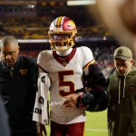 Ein Football-Spieler in einem weißen Washington-Trikot (#5), möglicherweise Jayden Daniels, mit einer großen schwarzen Armschiene (Hinweis auf Ellbogenverletzung), wird von zwei Mitarbeitern vom Spielfeld begleitet. Im Hintergrund sind Stadionsitze und das Spielfeld zu sehen. Diese Beschreibung wurde mit der FootballR KI automatisch generiert.