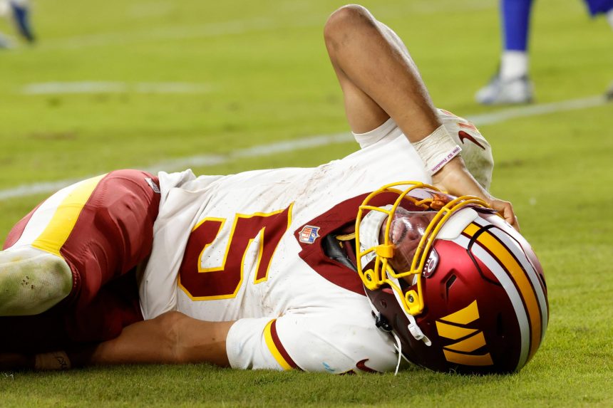 Ein Footballspieler in der Uniform der Washington Commanders liegt auf dem Spielfeld und hält sich vor Schmerzen den Kopf. Sein Helm bleibt auf, während er Grimassen schneidet; Grasflecken zieren seine kastanienbraune und goldene Uniform. Die Mannschaftskameraden, die in der Nähe stehen, warten darauf, dass Jayden Daniels über seine Verletzung berichtet. Diese Beschreibung wurde mit der FootballR KI automatisch generiert.