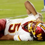 Ein Footballspieler in der Uniform der Washington Commanders liegt auf dem Spielfeld und hält sich vor Schmerzen den Kopf. Sein Helm bleibt auf, während er Grimassen schneidet; Grasflecken zieren seine kastanienbraune und goldene Uniform. Die Mannschaftskameraden, die in der Nähe stehen, warten darauf, dass Jayden Daniels über seine Verletzung berichtet. Diese Beschreibung wurde mit der FootballR KI automatisch generiert.