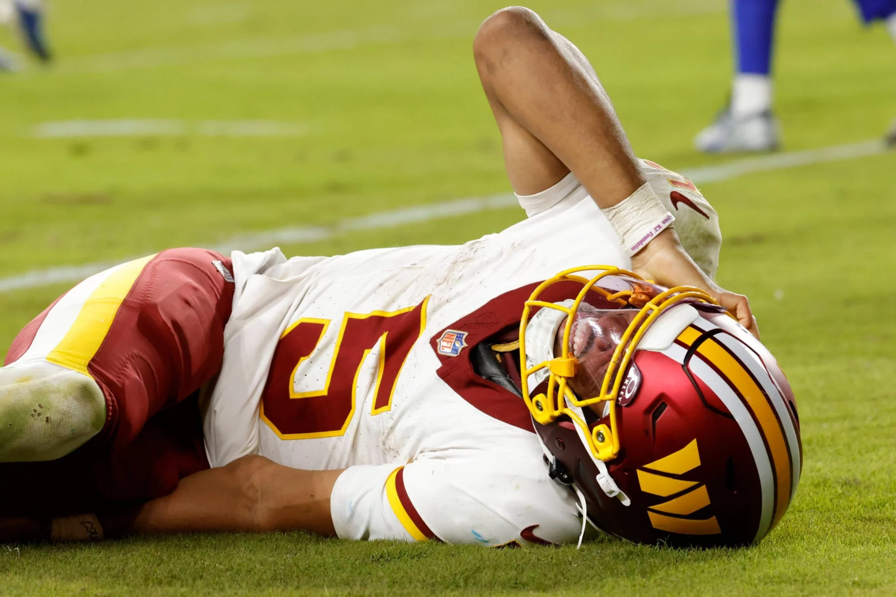 Ein Footballspieler in der Uniform der Washington Commanders liegt auf dem Spielfeld und hält sich vor Schmerzen den Kopf. Sein Helm bleibt auf, während er Grimassen schneidet; Grasflecken zieren seine kastanienbraune und goldene Uniform. Die Mannschaftskameraden, die in der Nähe stehen, warten darauf, dass Jayden Daniels über seine Verletzung berichtet. Diese Beschreibung wurde mit der FootballR KI automatisch generiert.