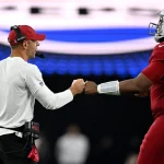 Ein Football-Trainer in einem weißen Hemd und einer roten Mütze gibt dem Cardinals-Spieler Nummer 7, Kyler Murray, in einer roten Arizona Cardinals-Uniform und einem roten Helm, einen Faustschlag. Der Hintergrund ist unscharf mit hellen Stadionlichtern und einem großen Schild über dem Kopf. Diese Beschreibung wurde mit der FootballR KI automatisch generiert.