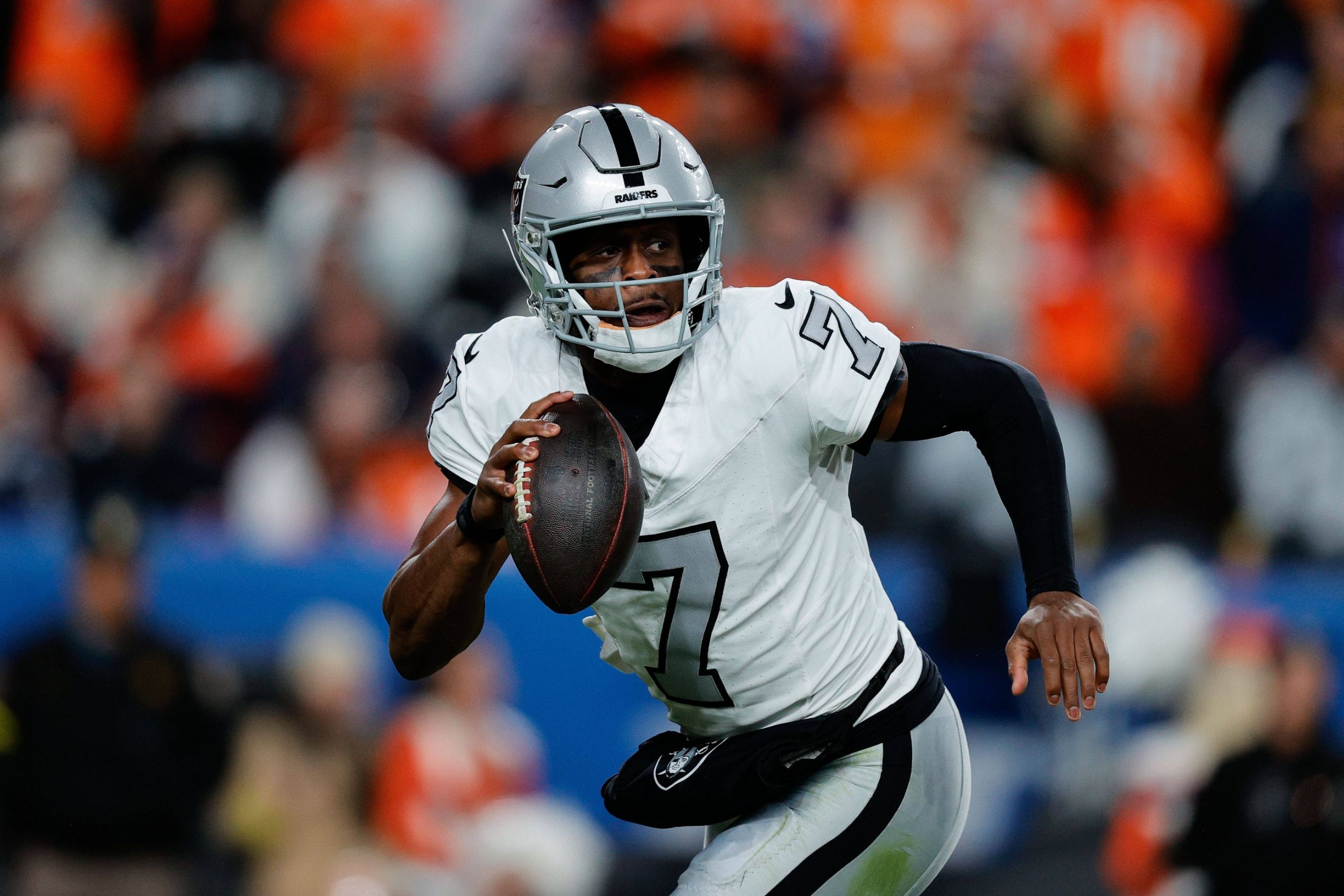 Quarterback Geno Smith - Nummer 7 - der Las Vegas Raiders läuft mit dem Ball in der rechten Hand, den Blick nach vorne gerichtet. Mit seinem silbernen Helm und den schwarzen Handschuhen stürmt er vorwärts, während die orange-blaue Menge im Hintergrund verschwimmt. Diese Beschreibung wurde mit der FootballR KI automatisch generiert.