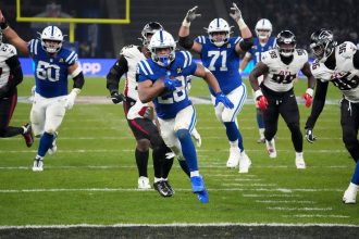 NFL Playoff Picture - Jonathan Taylor in einem blauen Trikot der Indianapolis Colts läuft mit dem Ball, verfolgt von Verteidigern der Atlanta Falcons in Weiß und Schwarz, während eines NFL-Playoff-Spiels. Mannschaftskameraden blockieren auf dem grünen Spielfeld; gelbe Torpfosten und eine unscharfe Menschenmenge füllen den Hintergrund. Diese Beschreibung wurde mit der FootballR KI automatisch generiert.