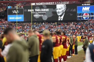 Die Zuschauer im Stadion beobachten, wie die Football-Spieler auf dem Feld stehen. Auf einer großen Leinwand wird eine Hommage an die NFL-Legende und den ehemaligen Commissioner Paul Tagliabue (1940-2025) gezeigt, um sein Vermächtnis während des Spiels zu würdigen. Diese Beschreibung wurde mit der FootballR KI automatisch generiert.