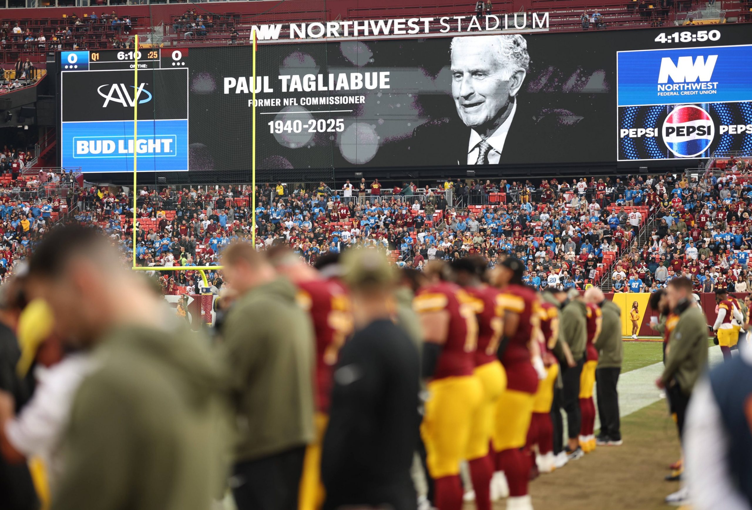 Die Zuschauer im Stadion beobachten, wie die Football-Spieler auf dem Feld stehen. Auf einer großen Leinwand wird eine Hommage an die NFL-Legende und den ehemaligen Commissioner Paul Tagliabue (1940-2025) gezeigt, um sein Vermächtnis während des Spiels zu würdigen. Diese Beschreibung wurde mit der FootballR KI automatisch generiert.