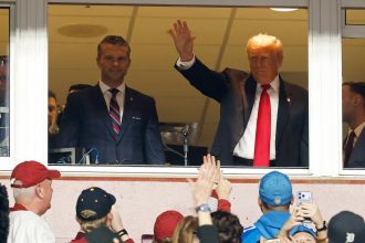 Zwei Männer stehen hinter einem Fenster über einer Menschenmenge. Der Mann rechts im dunklen Anzug und mit roter Krawatte, Donald Trump, winkt den Leuten unten zu. Einige im Vordergrund, vielleicht bei einem NFL-Spiel, schauen mit erhobenen Telefonen und Hüten in verschiedenen Farben auf. Diese Beschreibung wurde mit der FootballR KI automatisch generiert.
