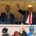 Zwei Männer stehen hinter einem Fenster über einer Menschenmenge. Der Mann rechts im dunklen Anzug und mit roter Krawatte, Donald Trump, winkt den Leuten unten zu. Einige im Vordergrund, vielleicht bei einem NFL-Spiel, schauen mit erhobenen Telefonen und Hüten in verschiedenen Farben auf. Diese Beschreibung wurde mit der FootballR KI automatisch generiert.